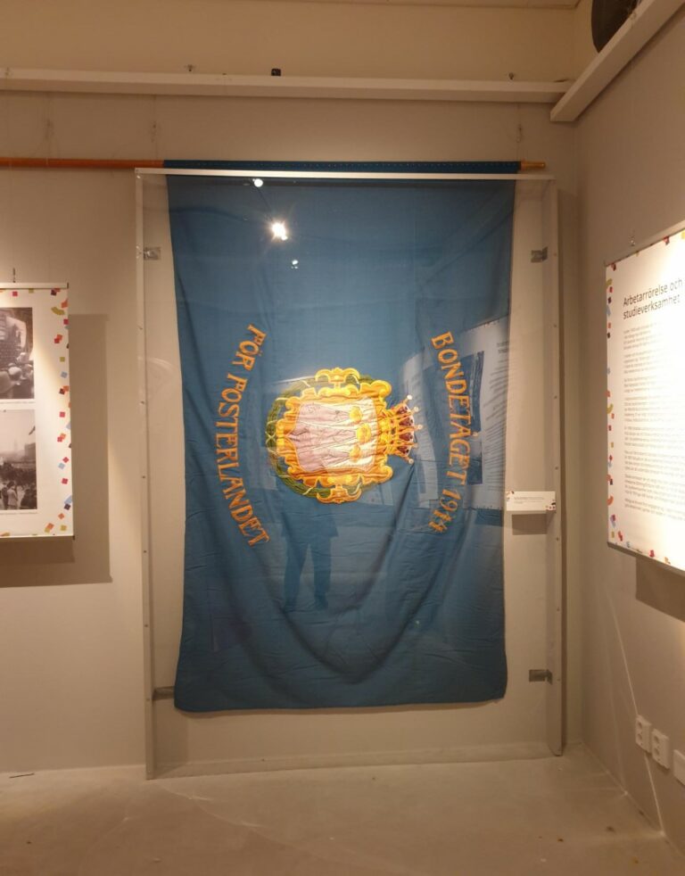 En blå fana i tyg hänger bakom ett glas på en vägg. I mitten på fanan stå det Bondetåget 1914 för fosterlandet. I mitten finns västmanlands landskapsvapen broderat.