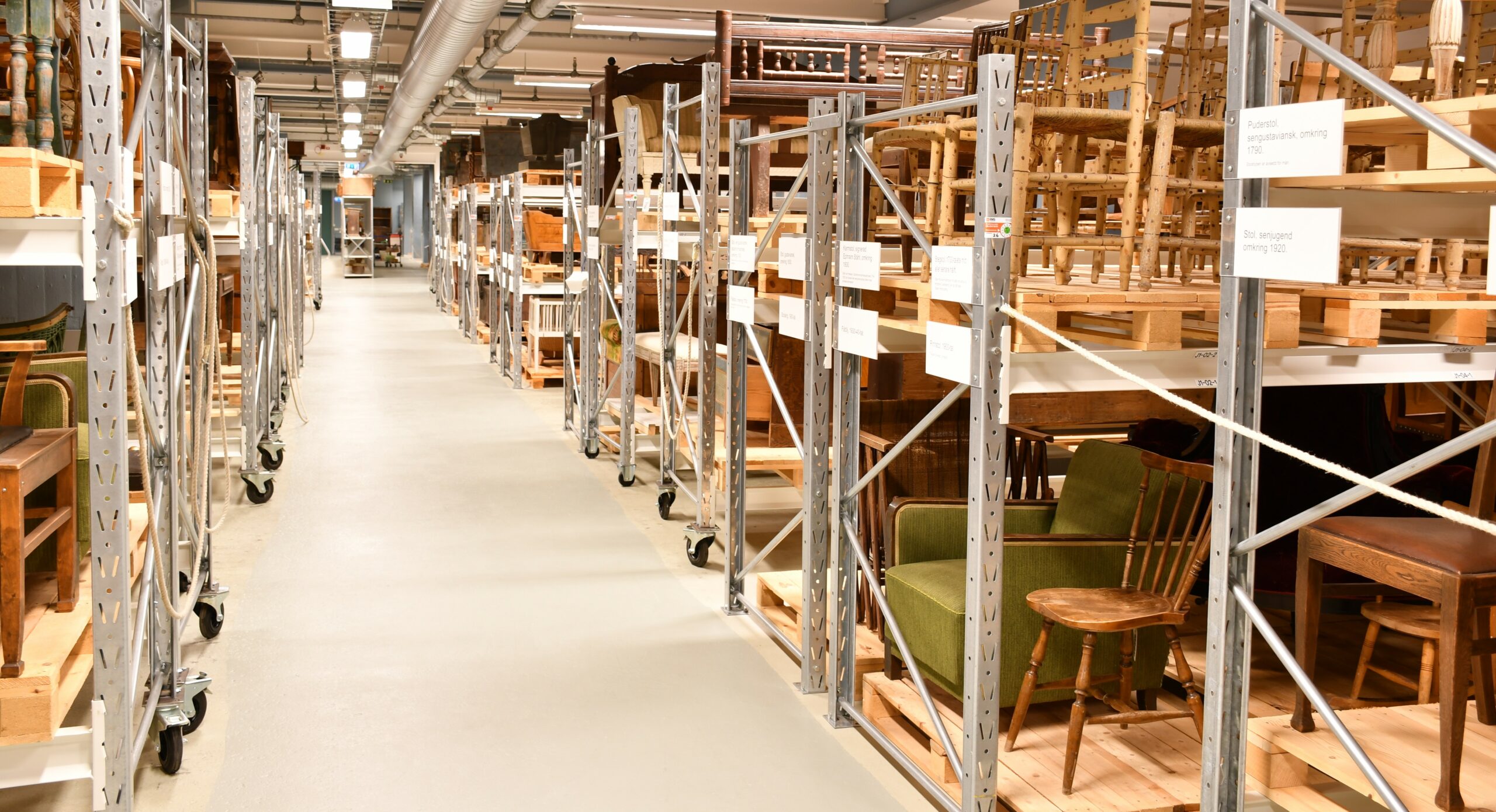 En överblick över lagret för föremålssamlingar. En lång korridor i mitten och bredvid står flera rader av metallhyllor där det står möbler.