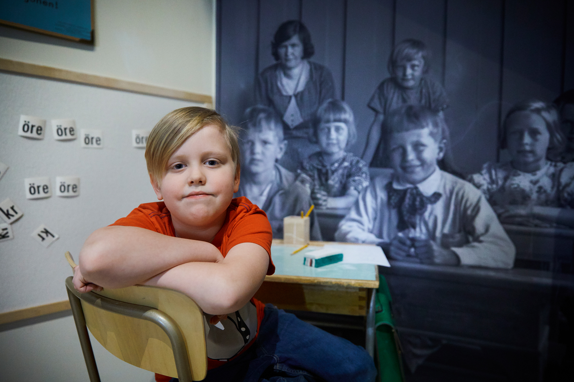 Färgfoto. Ett barn sitter vid en skolbänk i en museimiljö. Bakom barnet syns en stor svartvit bild av en äldre skolklass med barn och en lärare.