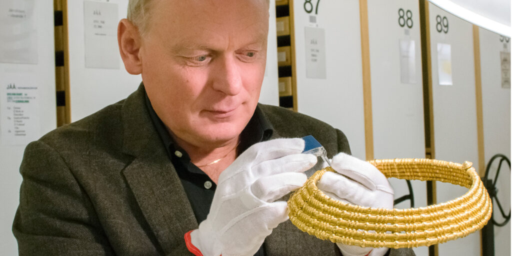 En man står och undersöker ett halsband i guld. Mannen har vita handskar på händerna och en brun kavaj. Bakom honom ser man en rad med vita plåtskåp.