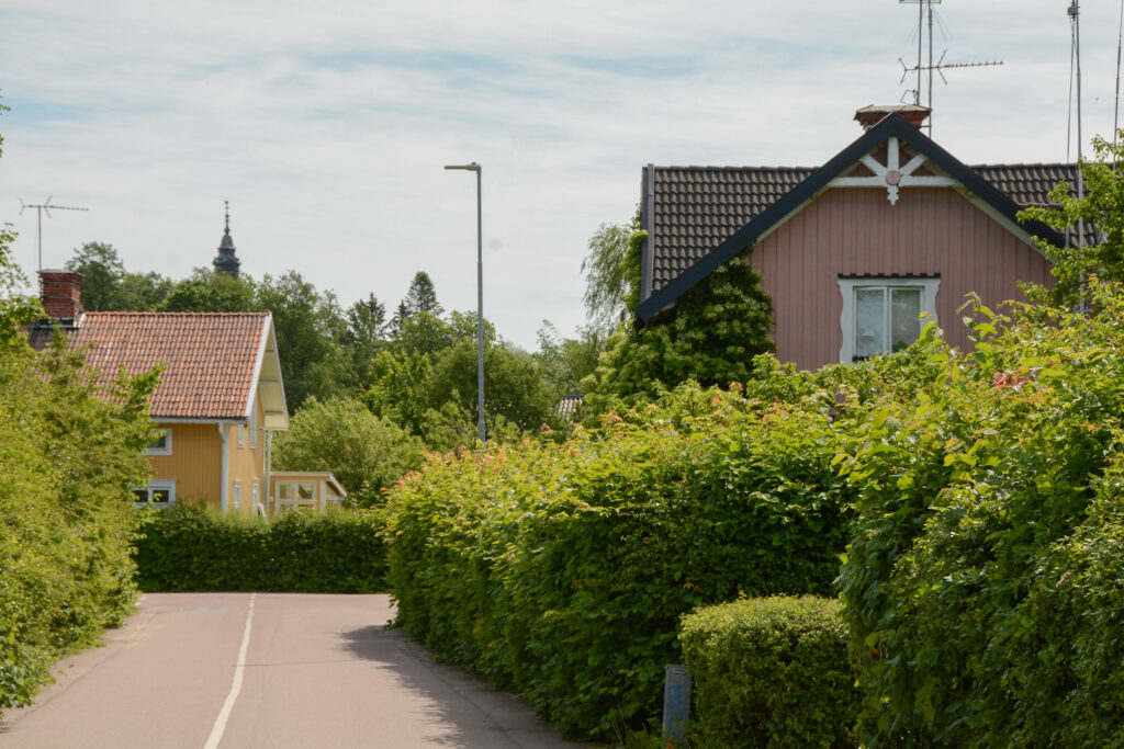 En sommarbild över ett villaområde, Man ser några rosa och gula träbyggnader och en massa gröna buskar längs en väg.