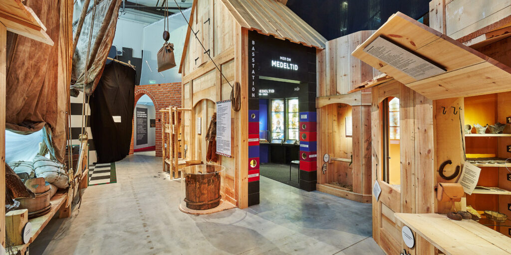 En översiktsbild från länsmuseets utställning om medeltiden. Man ser en uppbyggd hamnmiljö mestadels i trä. På höger sida ser man en handelsbod i trä.