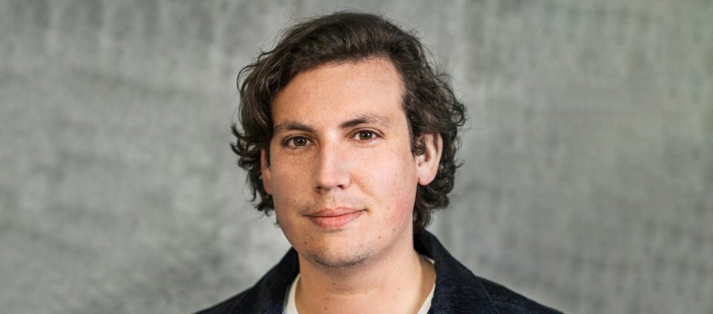 Färgfoto av en person med mörkt, lockigt hår som tittar rakt in i kameran. Bakgrunden är ljust grå och oskarp.