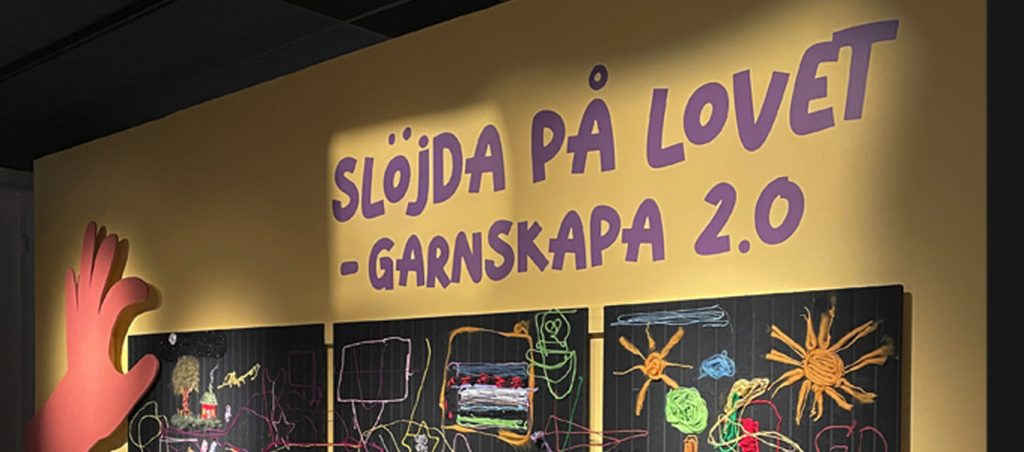 En gul vägg med lila texten ”Slöjda på lovet – garnskapa 2.0” och tre svarta tavlor nedanför, täckta av färgglada garnfigurer och mönster.