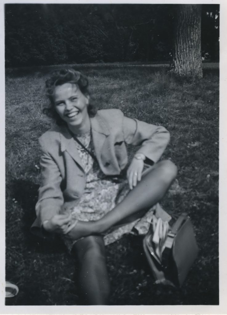 Svartvitt fotografi av en kvinna som sitter på gräset utomhus och skrattar medan hon håller i sin fot. En handväska ligger bredvid henne.