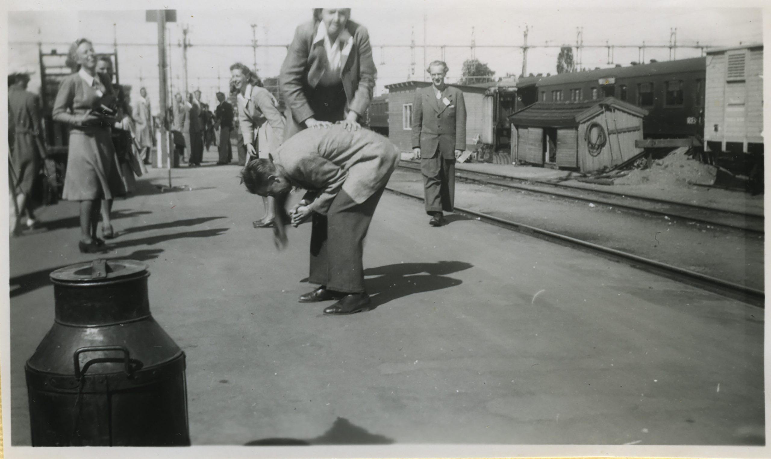 Svartvitt fotografi taget på en järnvägsperrong där flera personer samlats. I förgrunden håller en person en annan upp och ner, medan människor i bakgrunden ser på.