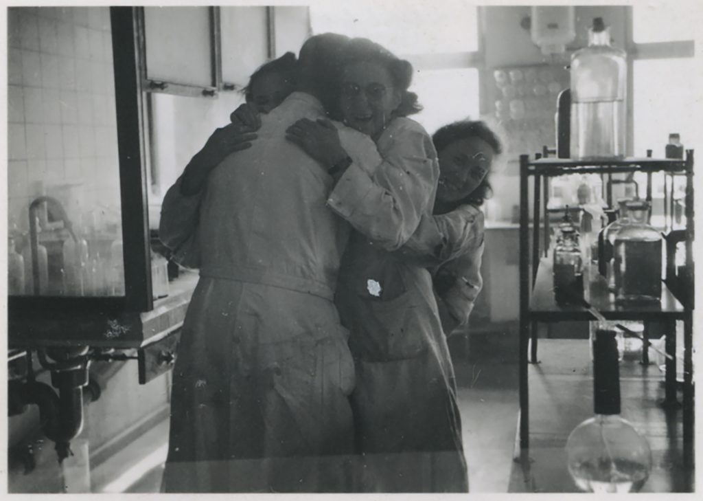 Svartvitt fotografi från ett laboratorium där tre personer i arbetsrockar kramar varandra. Laboratorieutrustning syns runtomkring.