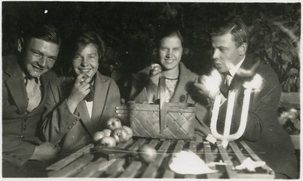 Svartvitt fotografi av fyra personer runt ett bord utomhus. De håller äpplen i händerna och en korg med äpplen står på bordet.