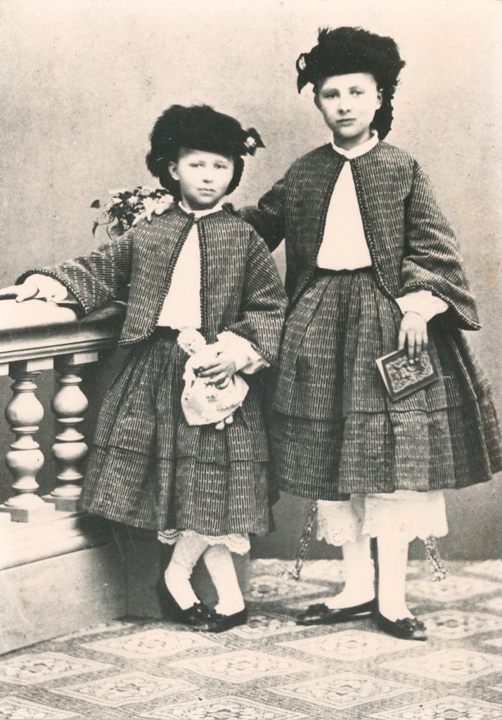 Svartvitt studiofotografi av två flickor som står uppställda bredvid ett bord. De bär likadana kläder och har allvarliga uttryck.