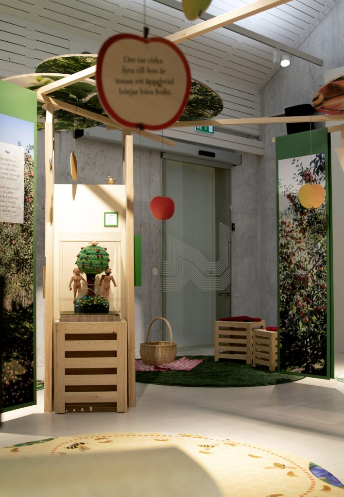 Färgfotografi. Översikt av en utställningsmiljö med träkonstruktioner, hängande äppelformade skyltar och dekor i grönt och rött. I mitten syns en glasmonter med ett äppelträd och två figurer. På golvet ligger en rund matta med bin, och intill finns korgar och sittlådor på en grön matta.