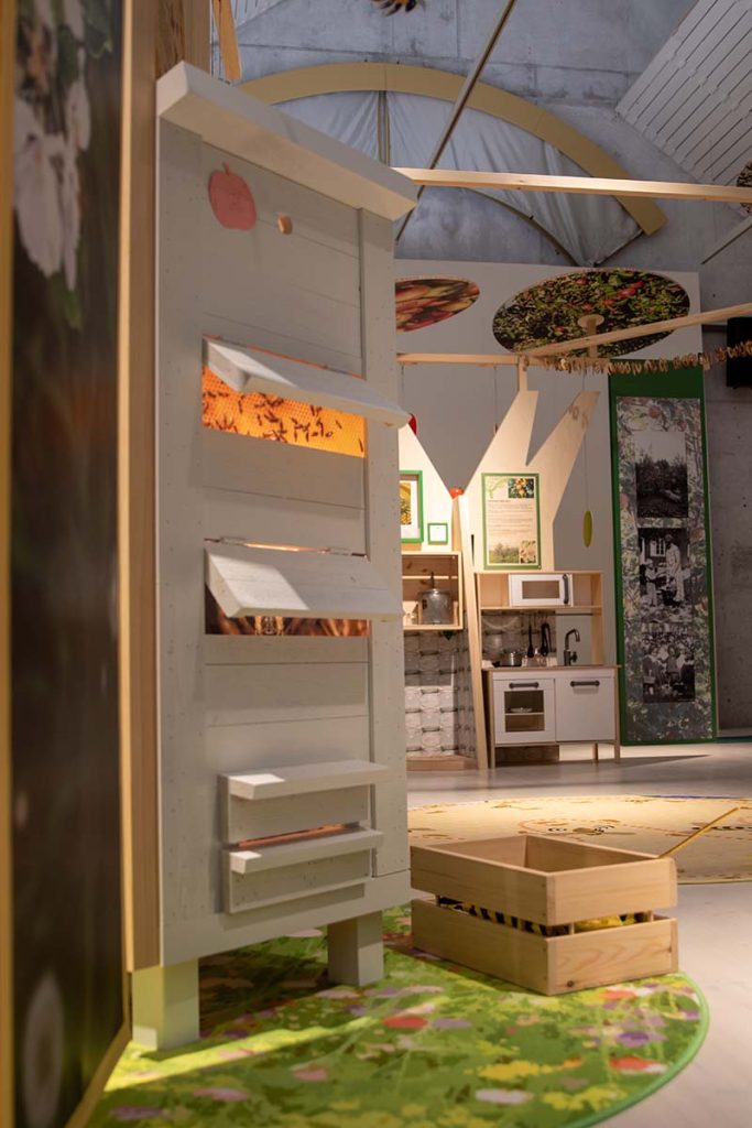 Färgfotografi. En vit bikupa i trä står i en utställningsmiljö. Genom öppningar syns bilder av bin. Trälådor och gröna mattor finns runtom.