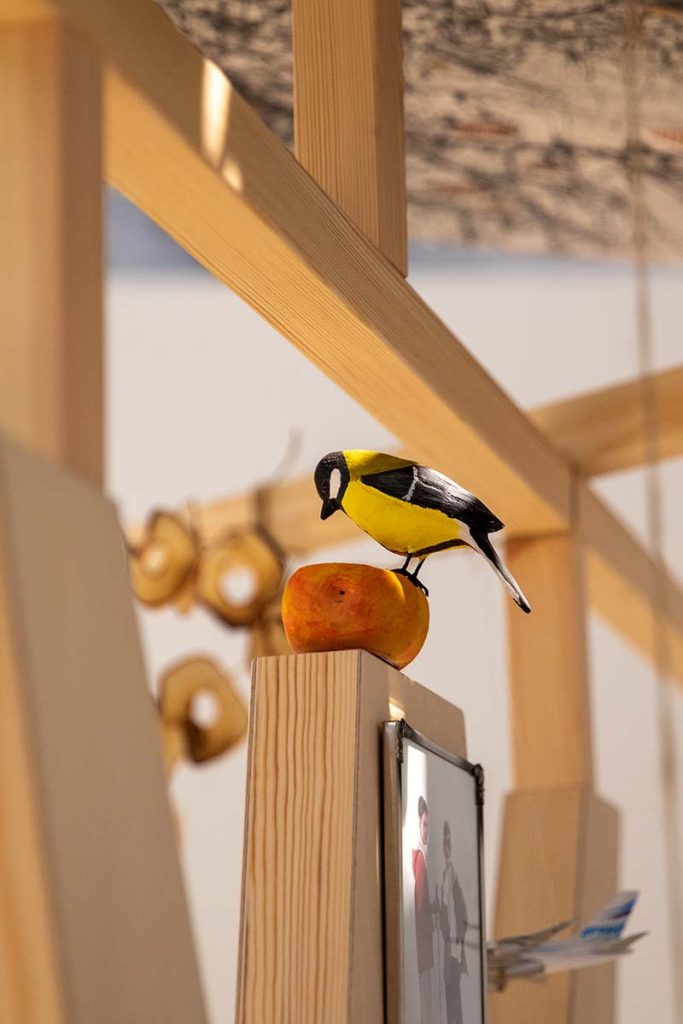 Färgfotografi. En liten fågelfigur sitter på ett äpple ovanpå en träställning i en utställning med ljusa träkonstruktioner i bakgrunden.