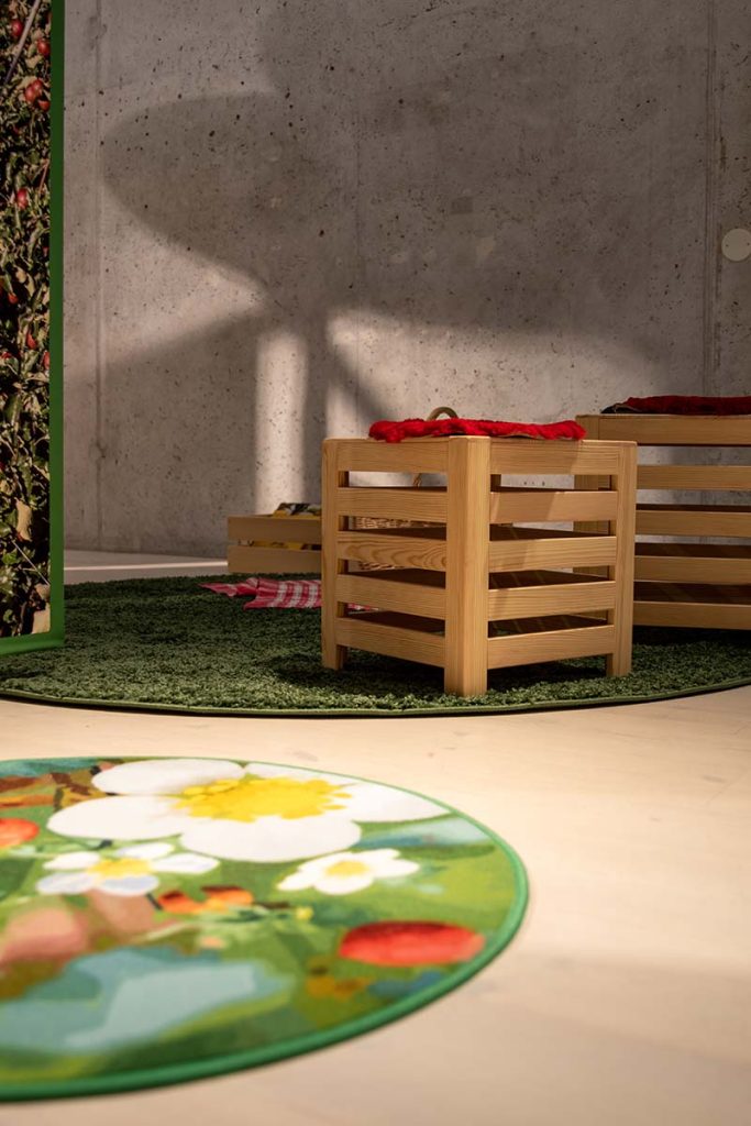 Färgfotografi. Trälådor står på en grön rund matta. I förgrunden syns en rund matta med blommor och naturmotiv.