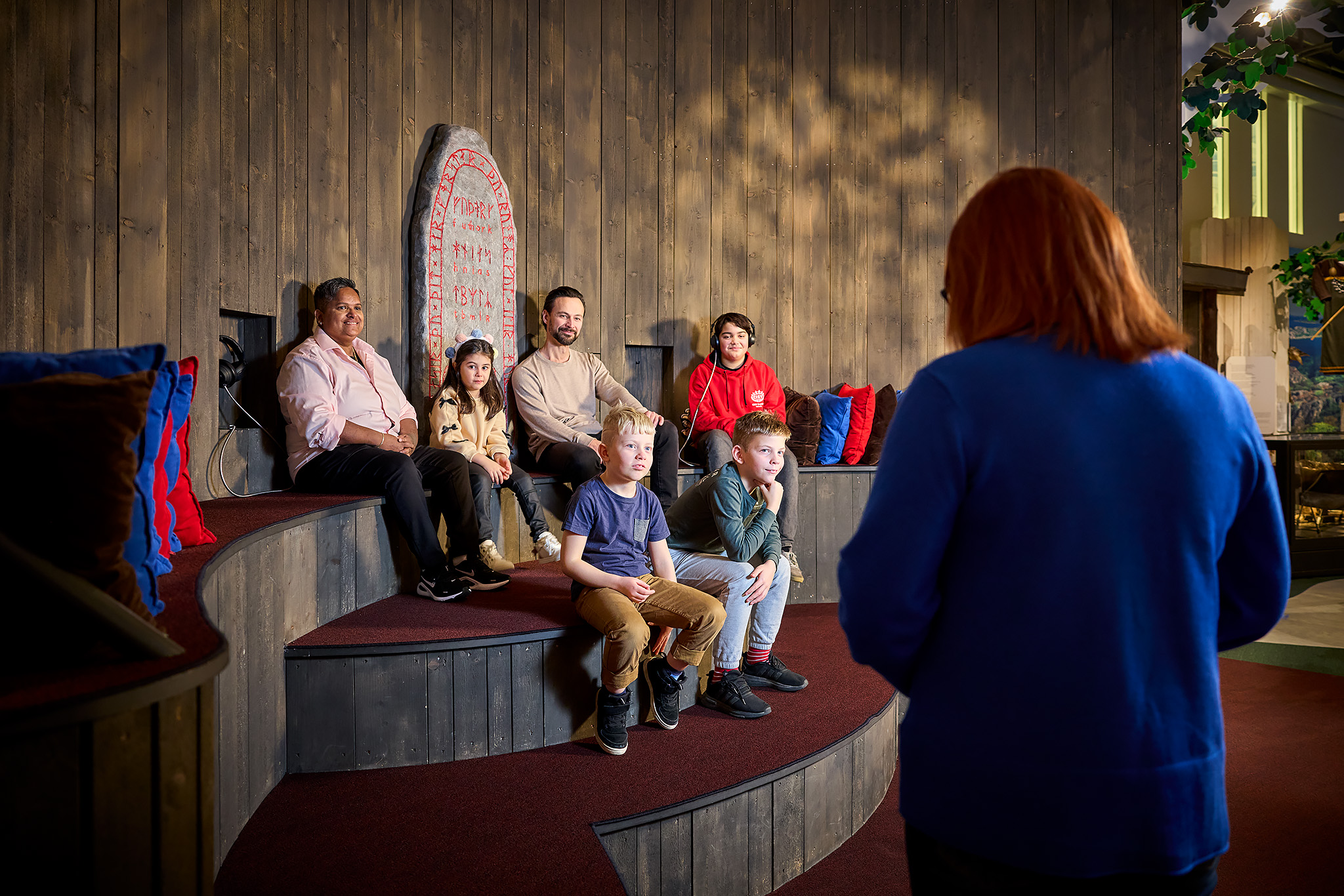 Färgfotografi. En grupp barn och vuxna sitter på en rund trägradäng i en museimiljö med träpanel och en runsten i bakgrunden. En person står framför gruppen och berättar.
