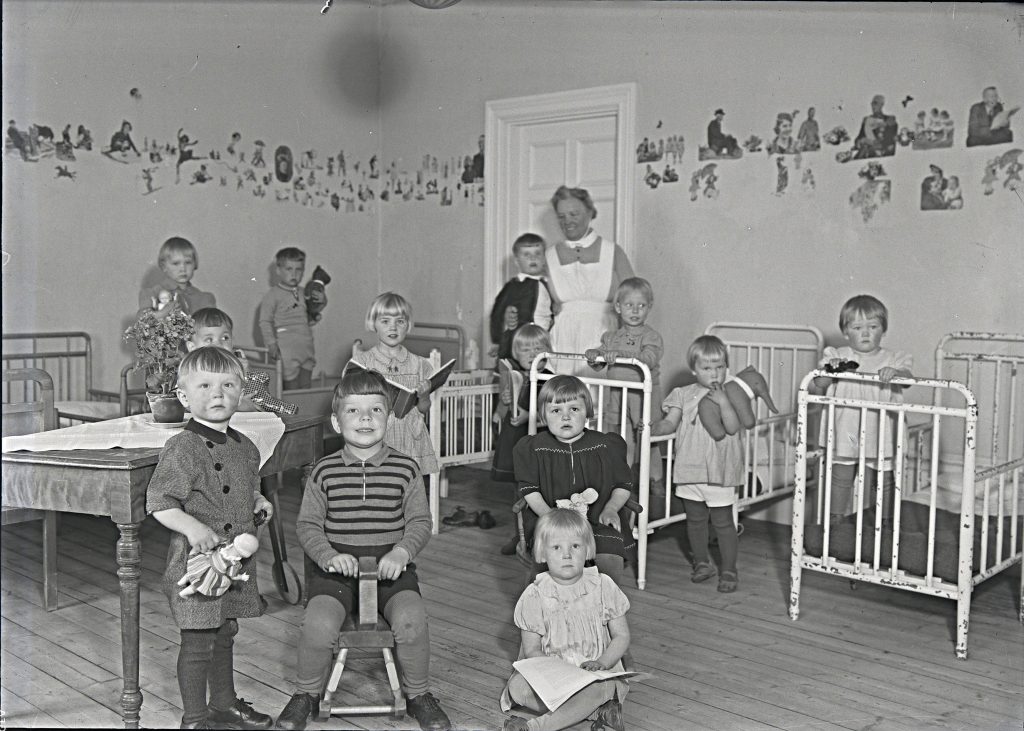 Svartvitt fotografi. En grupp små barn står och sitter i ett rum med järnsängar och leksaker. En vuxen kvinna i förkläde står bakom barnen.