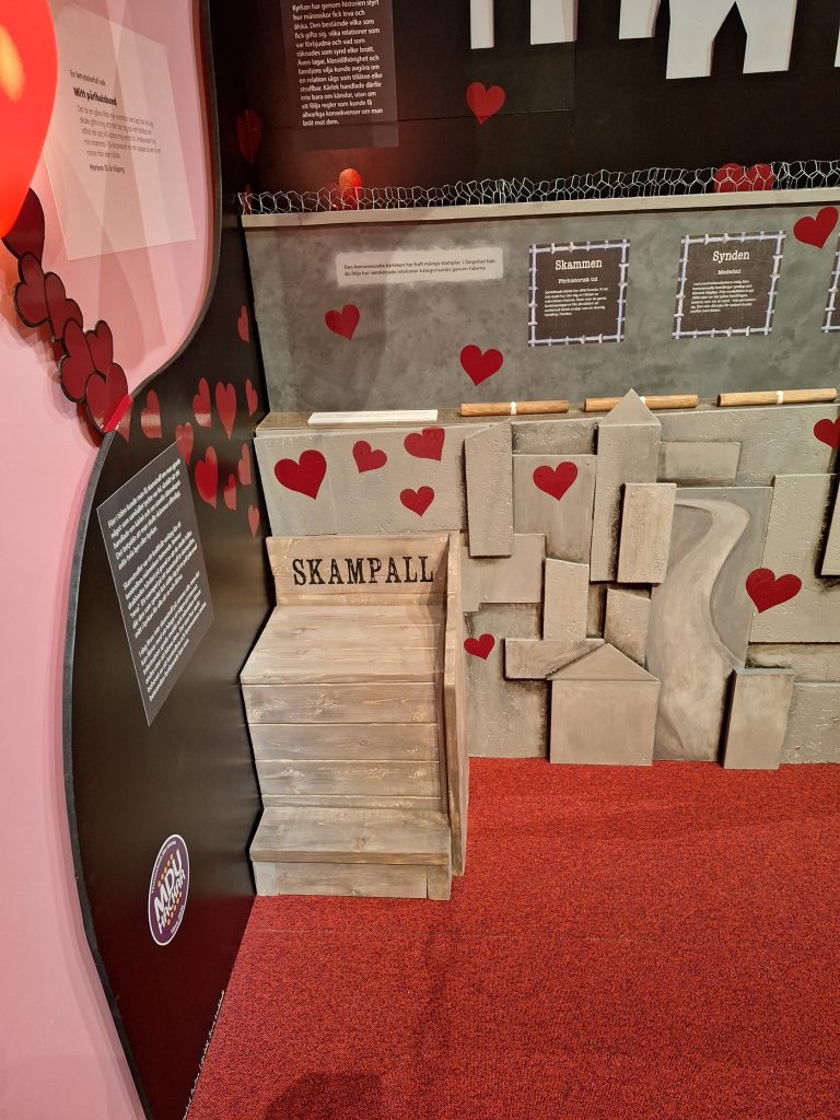 Färgfoto av utställningsdel med träkonstruktion märkt Skampall. Väggar dekorerade med röda hjärtan och texter om skam och synd.