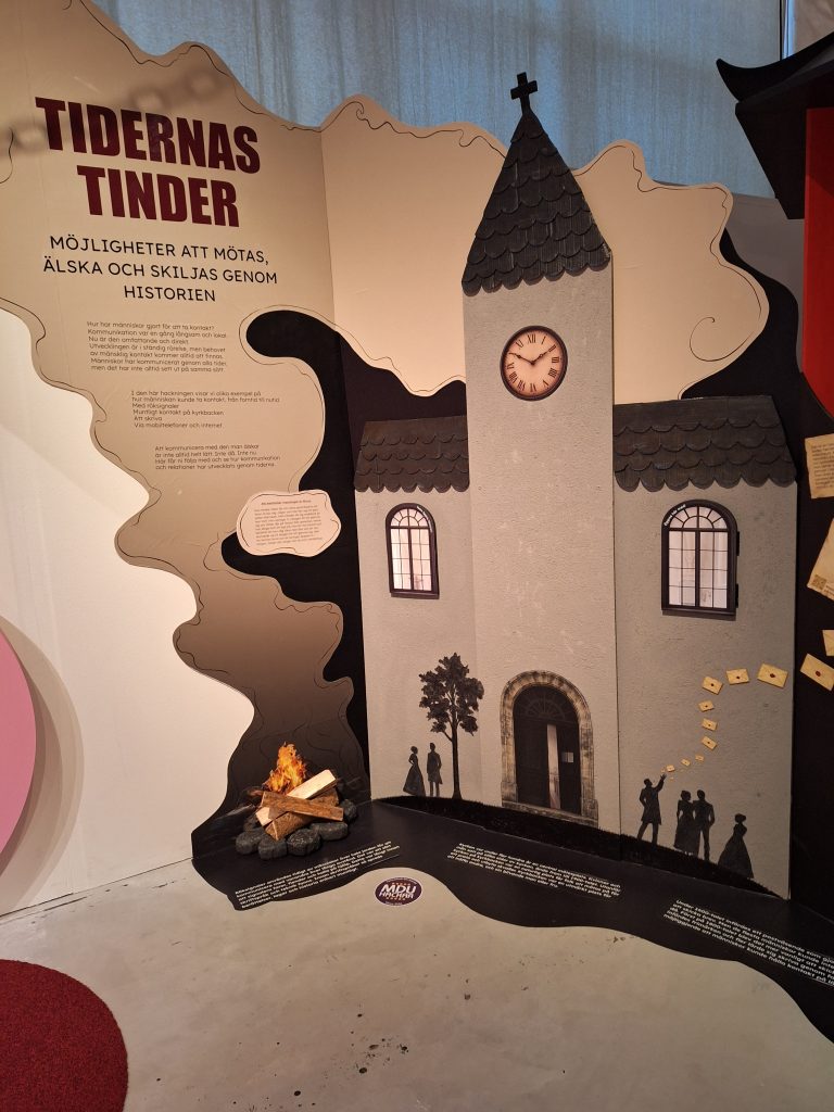 Färgfoto av utställningsdel med texten Tidernas Tinder. Illustration av kyrka, personer och eld som visar hur människor mötts genom historien.