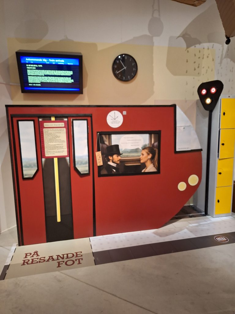 Färgfoto av utställningsdel formad som tågvagn med texten På resande fot. Inuti syns två personer som sitter mitt emot varandra.