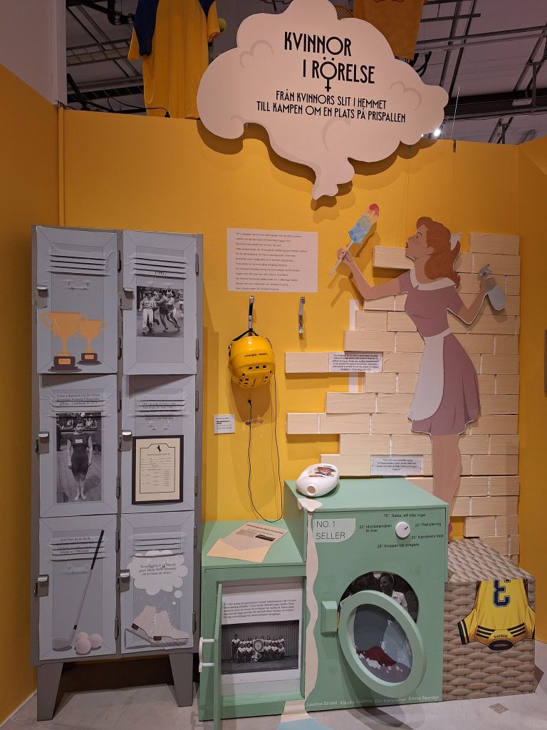 Färgfoto av utställningsdel med rubriken Kvinnor i rörelse. Skåp, idrottsföremål och illustration av kvinna som målar på en vägg.