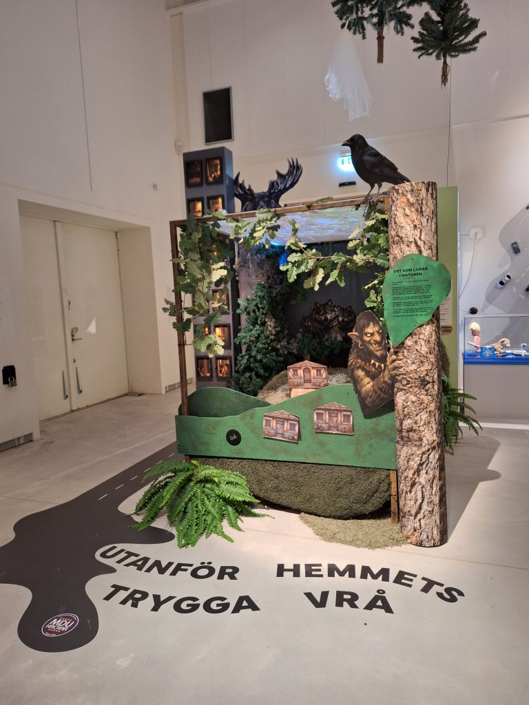 Färgfoto av utställningsdel med skogsmiljö. Träd, växter och figurer som föreställer djur och ett väsen.