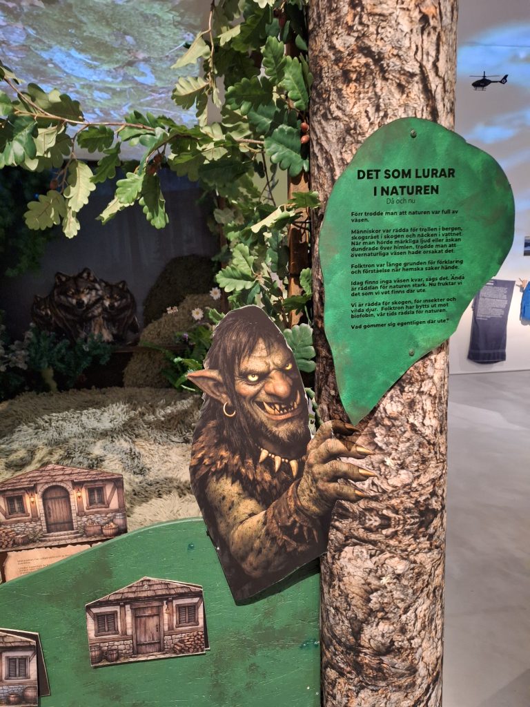 Färgfoto av detalj i utställning. Illustration av ett väsen som tittar fram bakom ett träd med text om naturens faror.