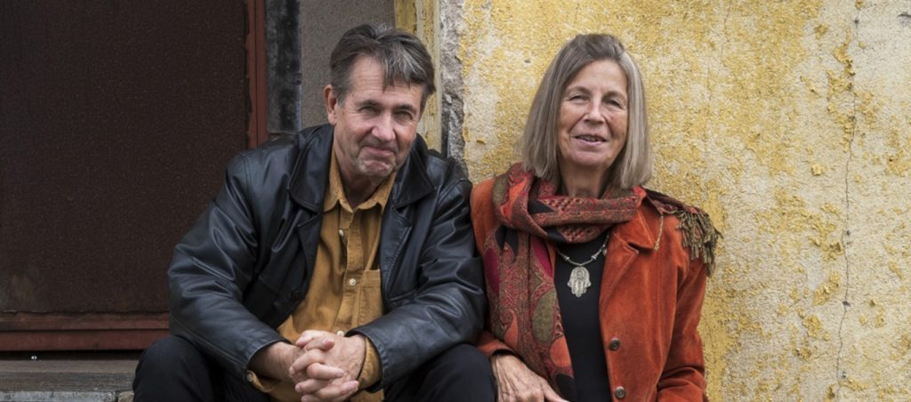 Färgfotografi. En man och en kvinna sitter bredvid varandra utanför en byggnad med gul putsad vägg och ser mot kameran.