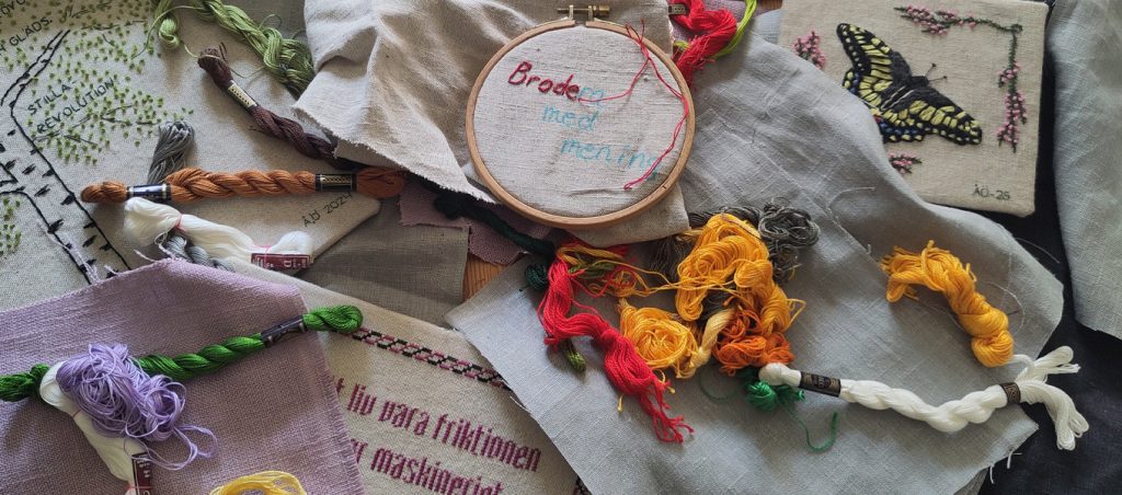 Broderimaterial utlagt på ett bord: färgglada garnhärvor, tygbitar och en broderiring med texten ”Brodera med mening”, omgiven av broderade motiv som en fjäril och växter.