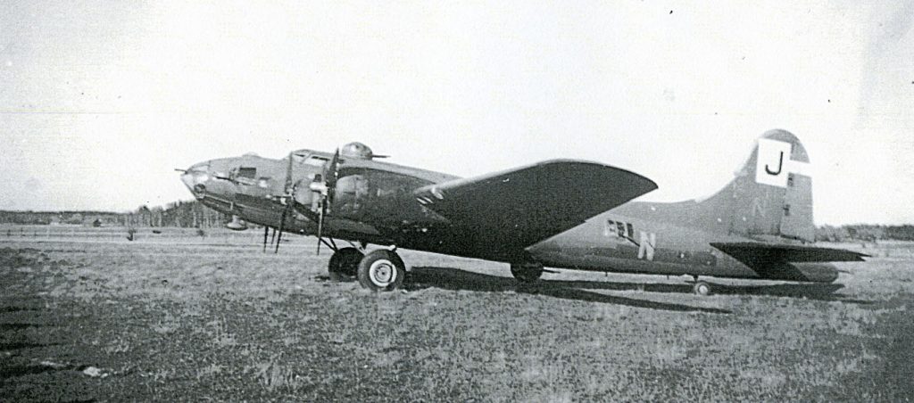 Svartvit bild av ett äldre militärt flygplan som står parkerat på ett öppet fält. Flygplanet har propellrar, rundad kropp och markerad stjärtfena med bokstaven “J”. I bakgrunden syns låg vegetation och en plan horisont.