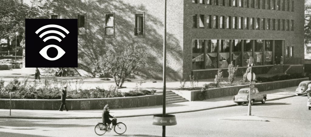 Svartvitt fotografi av stadsmiljö utanför Västerås stadsbibliotek. I förgrunden cyklar en person på en gata där även äldre bilar kör. På andra sidan gatan syns bibliotekets byggnad med stora fönster och människor som rör sig i området. I bildens övre vänstra hörn finns en symbol för syntolkning, en stiliserad ögonikon med ljudvågor.