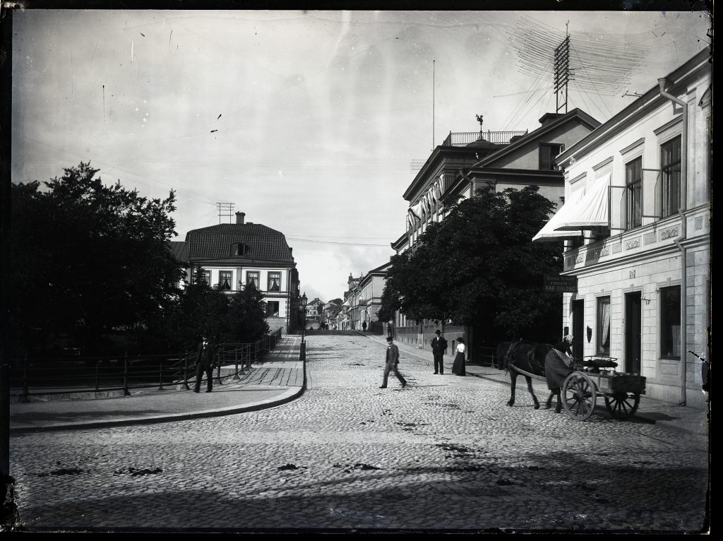 En häst med vagn står på en kullerstensgata framför stora stadsbyggnader. Några människor syns längs gatan.