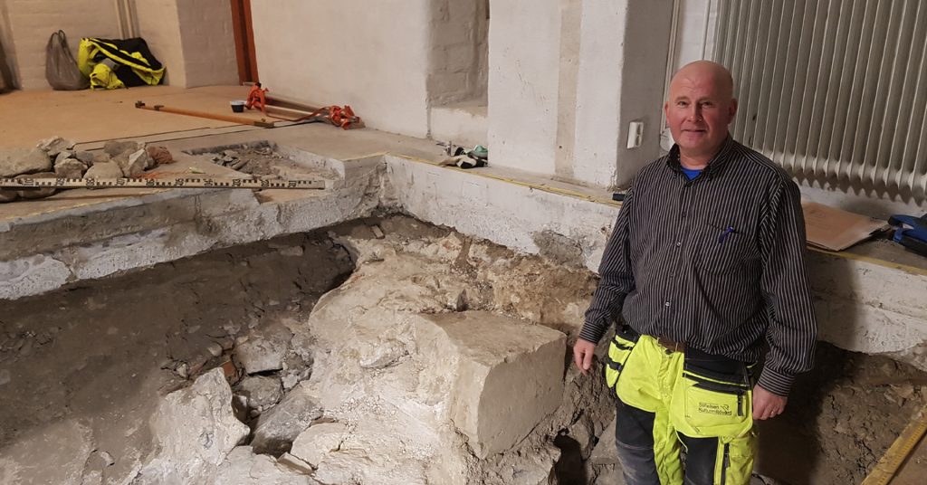 En man står bredvid en utgrävd yta i ett golv med stora stenar, jord och grus. I bakgrunden syns byggmaterial och utrustning.