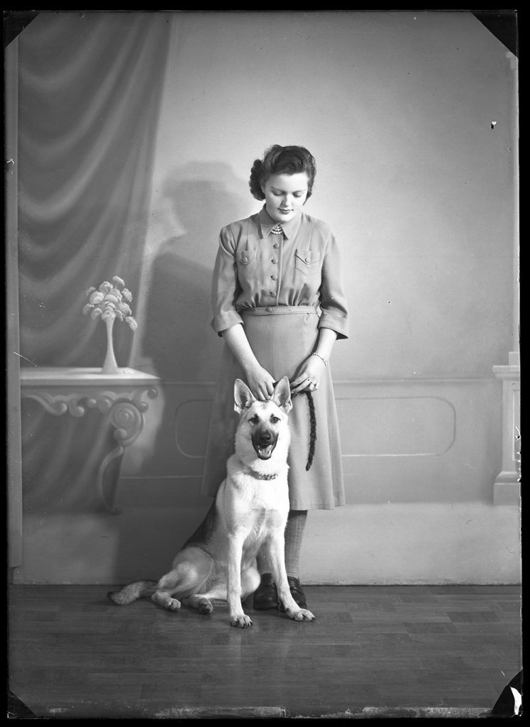 Svartvitt ateljéfotografi av en kvinna som står bakom en sittande hund och håller händerna vid hundens huvud.