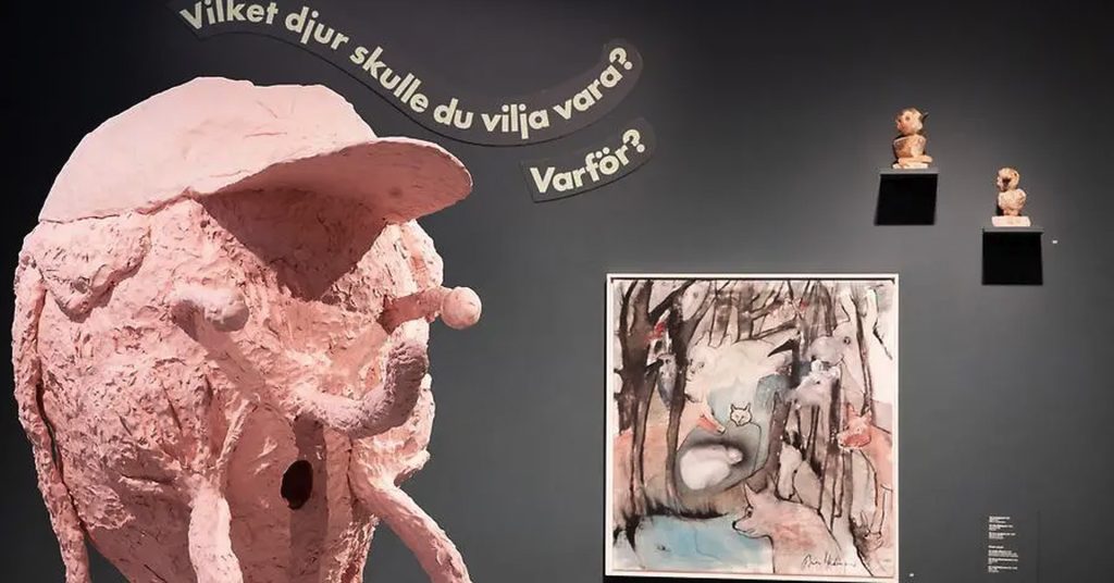Utställningsmiljö med en stor rosa skulptur till vänster, ett inramat konstverk på väggen och mindre skulpturer på svarta hyllor. På väggen står texten ”Vilket djur skulle du vilja vara? Varför?”.