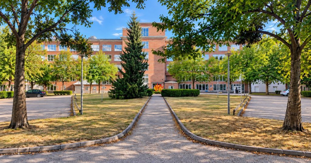 En gångväg leder genom en grönskande gård med stora träd och gräsytor fram till en tegelbyggnad. På båda sidor om gården står parkerade bilar.