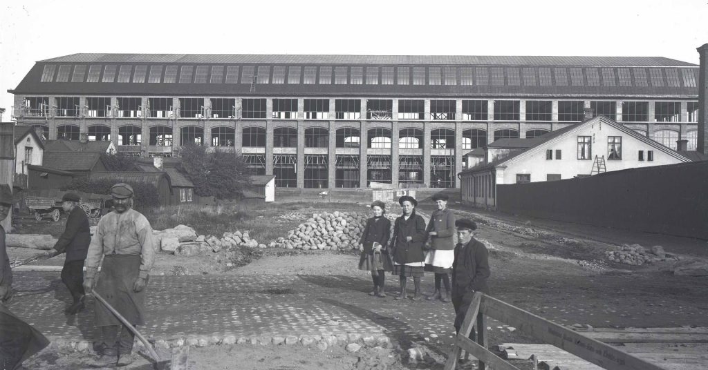 Svartvitt historiskt fotografi av en byggarbetsplats. I bakgrunden syns en stor industribyggnad och i förgrunden står flera personer bland sten, byggmaterial och en delvis stenlagd gata.