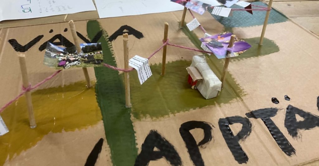 Närbild på ett modellbygge på kartong med målade gröna och gula ytor, träpinnar sammanbundna med rosa tråd samt små bilder och papperslappar som hänger längs tråden. I förgrunden syns stora svartmålade bokstäver på kartongen.